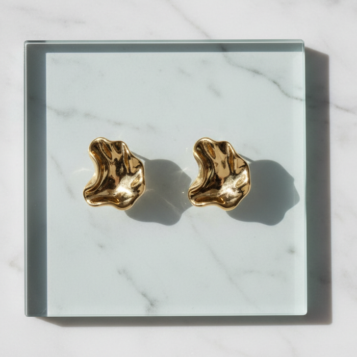 A pair of gold-toned, organic-shaped stud earrings displayed on a square glass tray over a white marble surface.
