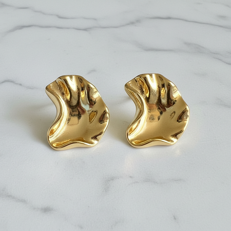 A pair of gold-toned, organic-shaped stud earrings with a polished, wavy surface, displayed on a white marble surface.
