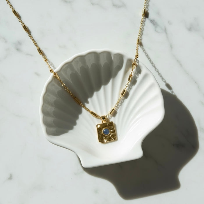 Gold necklace with a blue gemstone pendant on a white shell against a marble background