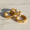 Three gold-toned textured rings with small blue gemstone accents, arranged on a light marble surface.