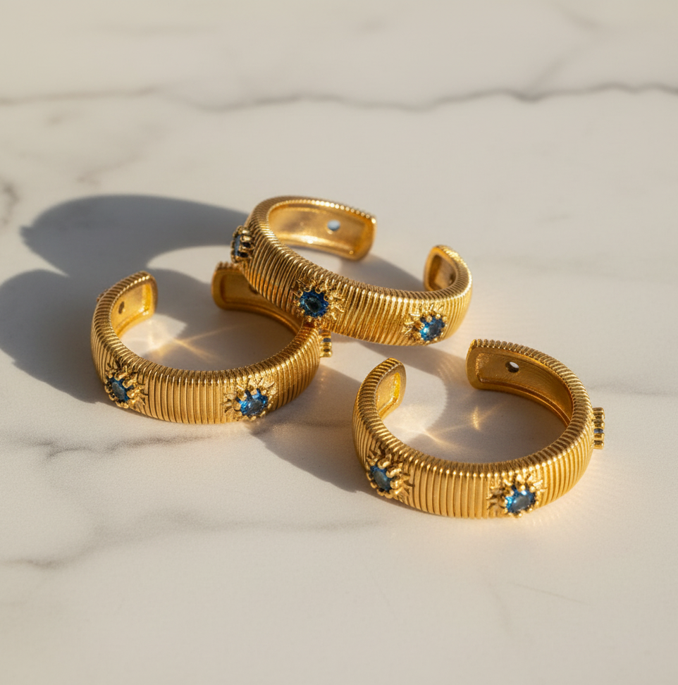 Three gold-toned textured rings with small blue gemstone accents, arranged on a light marble surface.