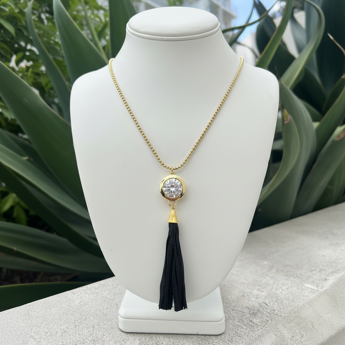 A gold chain necklace with a round bezel-set clear stone pendant and a long black tassel, displayed on a white jewelry bust with green foliage in the background.
