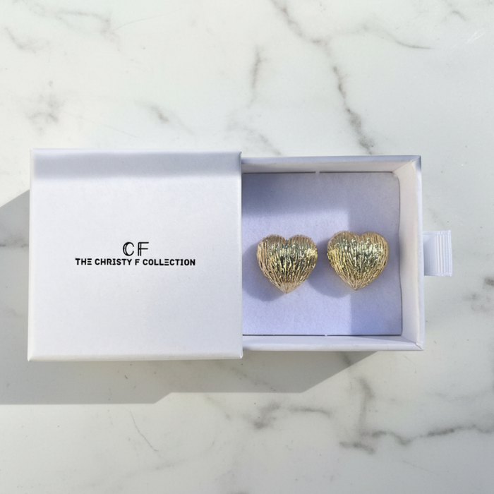 Gold heart-shaped earrings in a box with 'The Christy F Collection' branding on a marble surface.