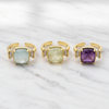 Three gold open ring earrings with cushion-cut gemstones in light blue, pale yellow, and purple, accented with small clear stones on a white marble background.