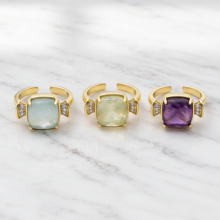 Three gold open ring earrings with cushion-cut gemstones in light blue, pale yellow, and purple, accented with small clear stones on a white marble background.