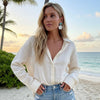 Woman in a white blouse and denim shorts standing on a beach with palm trees and ocean in the background.
