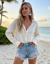 Woman in a white blouse and denim shorts standing on a beach with palm trees and ocean in the background.