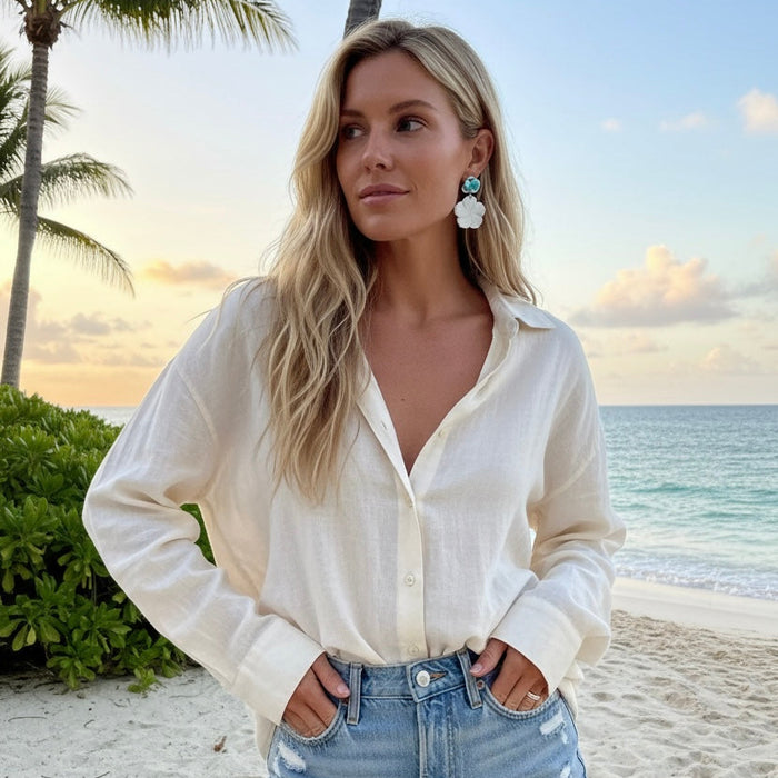 Woman in a white blouse and denim shorts standing on a beach with palm trees and ocean in the background.