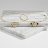 Two gold necklaces with small dangling gemstone charms, displayed on a white marble block against a light gray background.