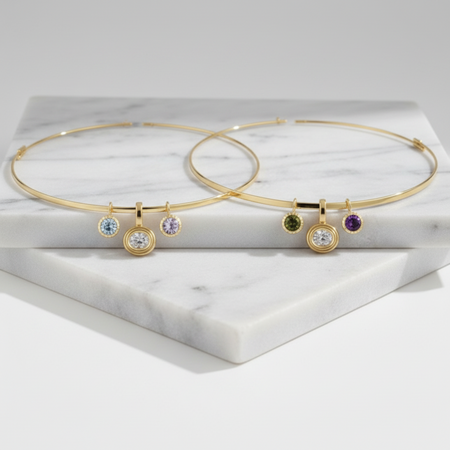 Two gold necklaces with small dangling gemstone charms, displayed on a white marble block against a light gray background.