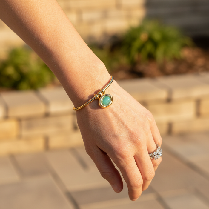 Hand wearing a gold bracelet with a green stone and a ring, outdoors.
