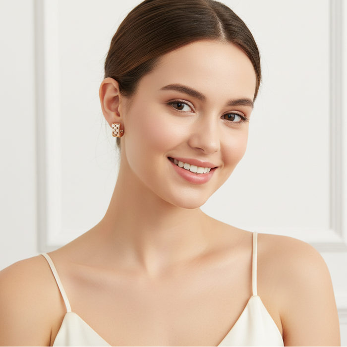 A smiling woman with her hair pulled back wears a small gold geometric square stud earring  photographed against a soft white paneled background.