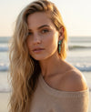 Woman with long blonde hair wearing a beige off-shoulder sweater on a beach.