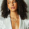 Woman with curly hair wearing a white blazer and layered necklaces against a neutral background