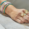 A close-up of a person's hand wearing multiple gold rings with oval pastel-colored stones and several stacked bangles in turquoise, lavender, and gold on the wrist, resting on a light marble surface.