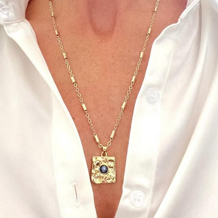A gold-filled necklace with a hammered pendant featuring a blue stone, worn by a person with a white shirt.
